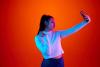 young lady showing peace gesture to camera while she takes selfie on smartphone against gradient red-orange background